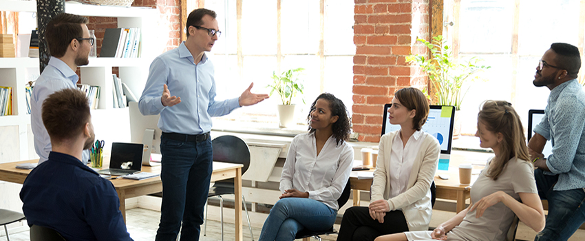 Man standing up talking with a group of people sitting and standing in an office listening.