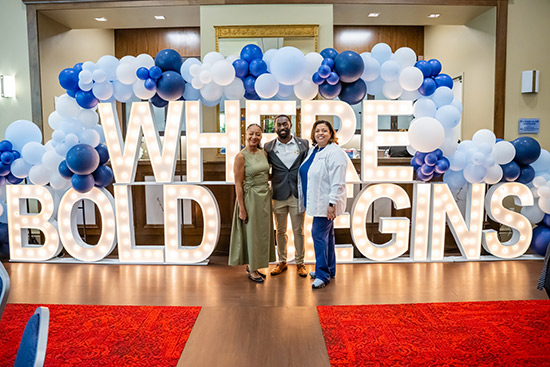Three people standing in front of the Where Bold Begins Campaign letter sign in the MacQueen Alumni Center.